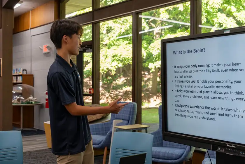 A presenter points to a large interactive display showing a slide titled “What is the Brain?” in a library room with tall windows